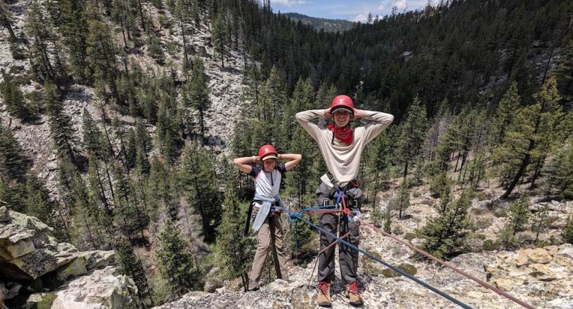 Two students wearing safety gear are secured by ropes as they lean back with their hands on their heads in a relaxed position.